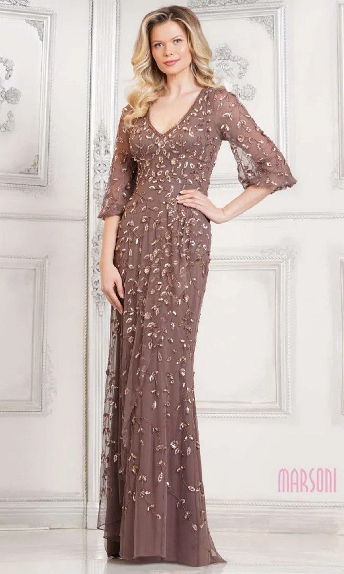 A woman stands before a decorative wall, wearing the Marsoni MV1320 long formal dress in dark taupe with sheer sleeves and floral embroidery—a refined, modest gown ideal for any elegant event.